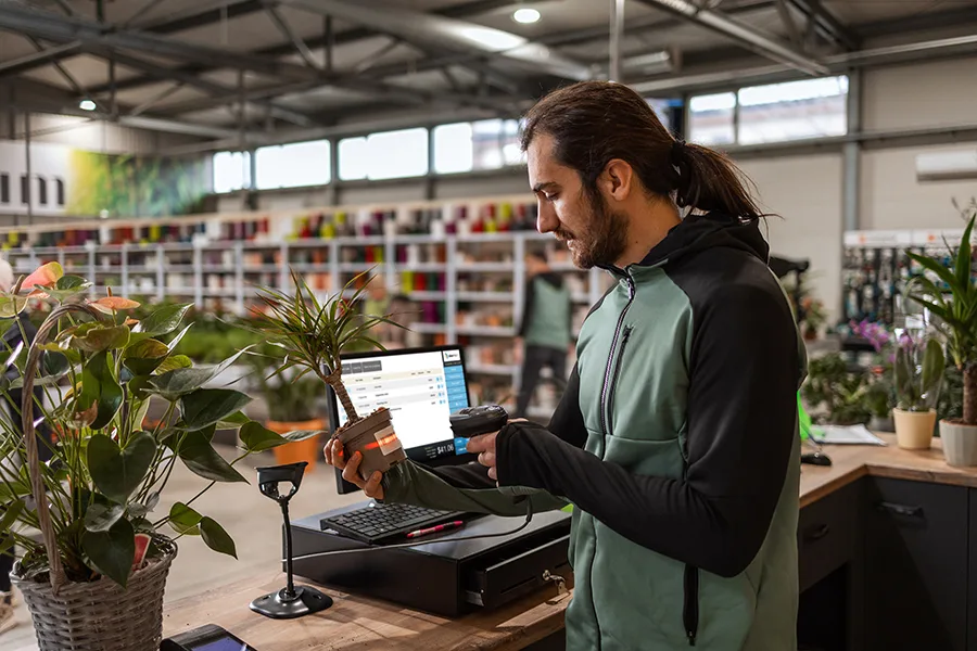 Employee scanning a plant at checkout using a Garden Center POS system inside a greenhouse retail store.